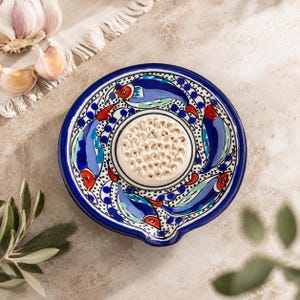 Peut inclure: Une assiette à râper le gingembre en céramique ronde avec une surface centrale surélevée et texturée pour râper, entourée d'un motif peint à la main de poissons bleus, rouges et vert clair sur fond blanc avec un bord bleu foncé et un bec verseur.