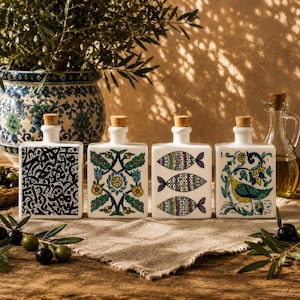 Peut inclure: Quatre bouteilles rectangulaires en céramique blanche avec des bouchons en liège, chacune présentant des motifs peints à la main : calligraphie noire, fleurs, trois poissons colorés et un oiseau parmi des fleurs. Les bouteilles sont alignées.