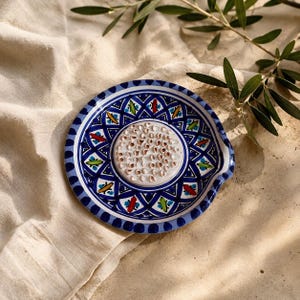 Peut inclure: Une assiette à râper ronde en céramique avec une surface centrale surélevée et texturée pour râper, entourée d'un motif géométrique bleu, blanc, vert et rouge. L'assiette présente une petite encoche sur le côté droit et une finition émaillée.