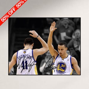 May include: A framed photograph featuring two basketball players in white Golden State Warriors jerseys with blue and yellow trim. One player wears number 11 with the name Thompson, and the other wears number 30. Both jerseys feature black ink autographs.