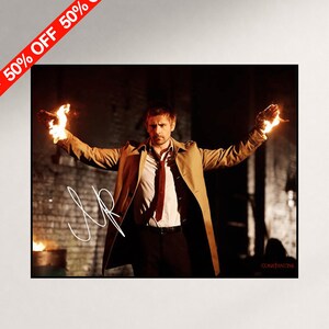 May include: A photographic print featuring a man in a tan trench coat, white shirt, and red tie, with both hands engulfed in flames. A white signature is overlaid on the left side, and the word Constantine is printed in small red text at the bottom right.