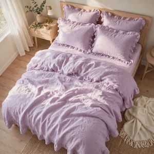 Puede incluir: Un juego de cama de color violeta claro que incluye una funda nórdica y cuatro fundas de almohada a juego, todas decoradas con bordes de volantes. La tela tiene una apariencia suave y texturizada, cubriendo una cama con cabecero de madera.