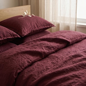 Peut inclure: Une parure de lit en lin bordeaux profond comprenant une housse de couette et deux taies d'oreiller assorties au tissage texturé visible. Le tissu présente un aspect naturel légèrement froissé, avec une branche d'eucalyptus séchée posée dessus.