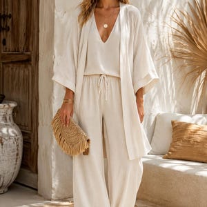 May include: A beige three-piece outfit consisting of a sleeveless V-neck top, wide-leg trousers with a drawstring waist, and an open-front kimono-style cardigan with wide sleeves. A tan woven clutch with fringe detail is held in one hand.