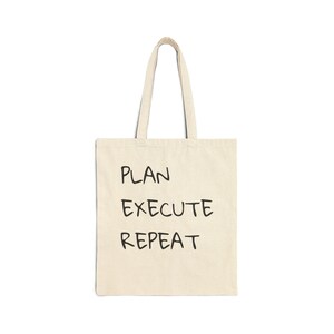 May include: A beige canvas tote bag with two shoulder straps, featuring the black text PLAN EXECUTE REPEAT printed in a simple, hand-drawn style font on the front center of the bag. The bag has a rectangular shape and a flat, open top design.