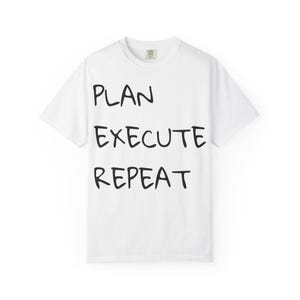 May include: A white short-sleeved t-shirt featuring the black text PLAN, EXECUTE, REPEAT printed in a hand-drawn style font on the front. The garment has a crew neckline and a standard fit, with a visible brand label inside the collar area.