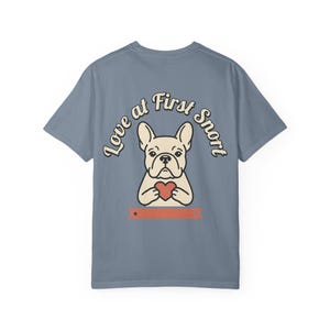 以下が含まれることがあります： スレートブルーの半袖Tシャツ。赤いハートを持ったクリーム色のフレンチブルドッグのグラフィックが描かれています。犬の上には「Love at First Snort」という文字がアーチ状に配置され、下部には2つの小さな星が付いた赤い水平のバナーがあります。