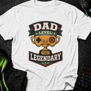 May include: A white t-shirt featuring a graphic print of a gold video game controller shaped like a trophy. The text reads DAD LEVEL: LEGENDARY and 1 PLAYER CHAMP. The design includes dark green banners, stars, and a small red heart icon.