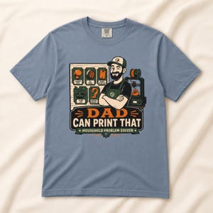May include: A dusty blue t-shirt featuring a graphic of a bearded person wearing a cap and apron, standing next to a 3D printer. The design includes icons of printed items and the text DAD CAN PRINT THAT HOUSEHOLD PROBLEM SOLVER.