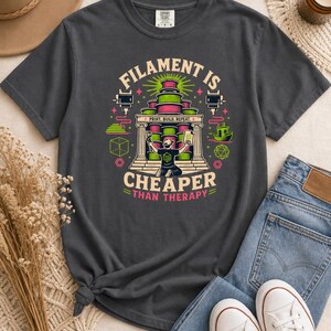 May include: A charcoal gray t-shirt featuring a colorful graphic of 3D printing spools, a printer nozzle, and a person holding a printed object. The text reads "FILAMENT IS CHEAPER THAN THERAPY" with "PRINT. BUILD. REPEAT." in the center.