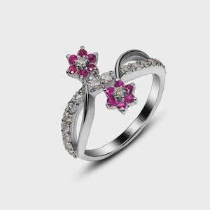 Könnte beinhalten: Ein silberfarbener Ring mit überkreuzter Ringschiene, der mit zwei blütenförmigen Elementen besetzt ist, die leuchtend rosa Steine und kleine klare Steine enthalten. Eine Seite der Ringschiene ist mit einer Reihe klarer Steine besetzt, was ein symmetrisches Design ergibt.