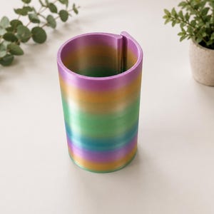 Könnte beinhalten: Ein zylindrischer Stiftehalter mit einer glatten Farbverlaufsoberfläche, die durch horizontale Bänder in Violett, Gold, Grün und Blau verläuft. Der Behälter hat eine vertikale Naht an einer Seite und bildet eine durchgehende, offene Struktur.