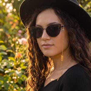 Op de afbeelding: Een zwarte fedora hoed en zwarte cat-eye zonnebril met een goudkleurige metalen ketting. De ketting is aan beide kanten voorzien van het jaartal 2023 in gouden cijfers en verbonden met het montuur. Gedragen met een zwart shirt met lange mouwen.