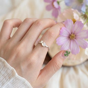 Puede incluir: Un anillo de plata con una pequeña piedra redonda e iridiscente engastada en una banda delicada. La banda tiene un acabado metálico pulido y un perfil delgado, diseñado para ajustarse cómodamente al dedo. El anillo es sencillo.