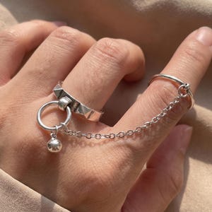 Può includere: Un doppio anello color argento collegato da una catenina sottile. Il primo anello presenta una fascia geometrica testurizzata con un ciondolo circolare pendente e una piccola sfera. Il secondo anello è una fascia liscia, collegata al primo da una catena.