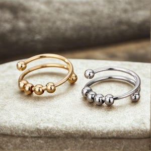 May include: Two open-band rings, one in a gold tone and one in a silver tone. Each ring features a double-wire design with a series of small, smooth, movable metal beads along the front section, allowing for tactile movement on the finger.