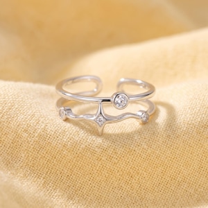 Puede incluir: Un anillo abierto de tono plateado con diseño de doble banda. La banda inferior tiene una forma ondulada con un motivo central de estrella de cuatro puntas y dos pequeñas piedras redondas transparentes. La banda superior incluye una piedra.