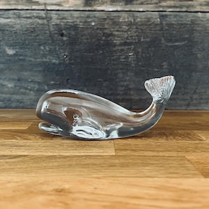 May include: A clear glass whale ornament with a detailed tail fin. The whale is placed on a wooden surface, with a rustic wooden backdrop. The ornament is a decorative item, suitable for a home or office.