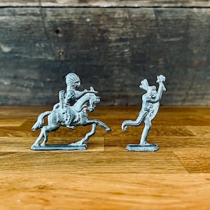 May include: Two small, silver-coloured metal toy figurines. One depicts a Native American riding a horse, the other a person running with a weapon. The figurines are on a wooden surface with a wooden background.