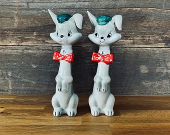 May include: Two ceramic bunny figurines with long necks, wearing red bow ties with white polka dots and green hats. The bunnies are standing on a wooden surface.