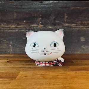 Puede incluir: Figura de gato de cerámica blanca con detalles en negro y rosa. El gato tiene ojos verdes, boca roja y bufanda a cuadros. La figura está sobre una superficie de madera.