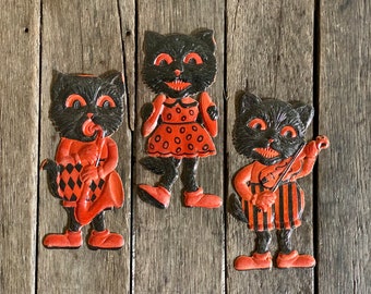 Könnte beinhalten: Drei schwarze Halloween-Katzen-Ausschnitte aus Papier mit orangefarbenen und schwarzen Details. Die Katzen spielen Musikinstrumente: ein Saxophon, eine Geige und eine Trommel.