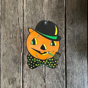 May include: A vintage Halloween decoration featuring an orange pumpkin head wearing a black hat with a green band and a black bow tie with green and yellow polka dots. The pumpkin is smoking a pipe.