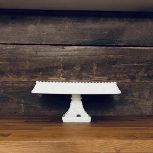 Puede incluir: Un soporte para pasteles rectangular blanco con un borde festoneado. El soporte está sostenido por una base de pedestal con detalles florales. Está hecho de un material opaco y sólido y se encuentra sobre una superficie de madera.