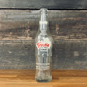 May include: A vintage Frostie soda bottle, clear glass with a red and white label. The label features the word "Frostie" in red script and the text "10 fl oz". The bottle is empty and has a classic design.