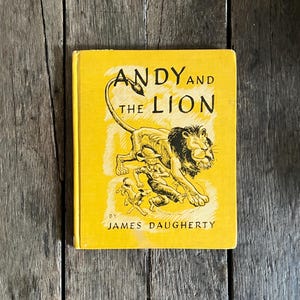 May include: A vintage children's book titled "Andy and the Lion" by James Daugherty. The book has a yellow cover with black text and illustrations of a lion and a boy. The book is set against a wooden background.