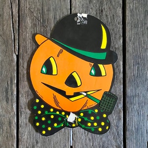 Könnte beinhalten: Eine Vintage-Halloween-Dekoration aus einem Kürbis, der einen schwarzen Zylinder mit grünem Band, eine grüne und gelbe gepunktete Fliege und eine Pfeife trägt. Der Kürbis hat ein gelbes und orangefarbenes Gesicht mit einem breiten Lächeln und grünen Augen. Der Text "Made in U.S.A." und "Beistle Co." ist auf der Dekoration aufgedruckt.