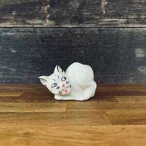Może przedstawiać: Biała ceramiczna figurka kota z zielonymi oczami, czarnymi oznaczeniami i czerwonymi ustami. Kot jest zwinięty, z głową obróconą na bok. Figurka stoi na drewnianej powierzchni.
