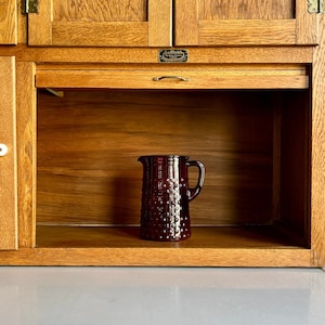 Op de afbeelding: Een donkerrode, getextureerde kan met een handvat staat in een houten kast. De kast heeft een roldeurplank en messing hardware. De houtnerf is zichtbaar op de kastdeuren en binnenkant.