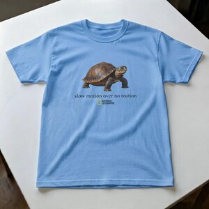 Puede incluir: Una camiseta de manga corta de color azul claro con un gráfico de una tortuga marrón en el centro. Debajo de la tortuga, el texto dice "slow motion over no motion" seguido del logotipo de National Geographic en su característico rectángulo amarillo.