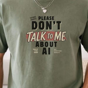 以下が含まれることがあります： オリーブグリーンのクルーネックTシャツ。前面には黒と赤の文字で「PLEASE DON'T TALK TO ME ABOUT AI」とプリントされ、その周囲に「NO MORE PROMPTS」「MADE BY A REAL HUMAN」「NOT EVERYTHING NEEDS AI」という小さな文字が添えられています。
