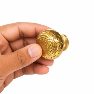 Könnte beinhalten: Ein runder Schrankknopf aus Messing mit einer strukturierten Oberfläche, einem gehämmerten Mittelteil und einem Rand aus geprägten kreisförmigen Motiven. Das Beschlagteil hat eine polierte Metalloberfläche und einen zylindrischen Sockel.