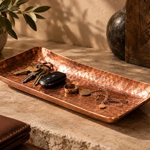 Peut inclure: Un plateau rectangulaire en cuivre martelé avec une finition métallique. Le plateau contient un trousseau de clés en métal avec un porte-clés noir, une bague en métal doré simple et un collier à chaîne dorée fine avec un petit pendentif circulaire.