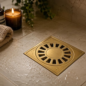 Peut inclure: Une grille de douche carrée en métal doré brossé, dotée d'une plaque centrale circulaire entourée d'une série de fentes de drainage rectangulaires, installée à fleur dans des carreaux de sol en pierre claire parsemés de gouttes d'eau.