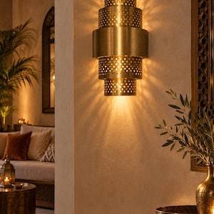 May include: A tiered brass wall sconce featuring a cylindrical design with alternating solid and perforated sections. The light fitting emits a warm glow, casting distinct light patterns onto the wall through the small circular cutouts.