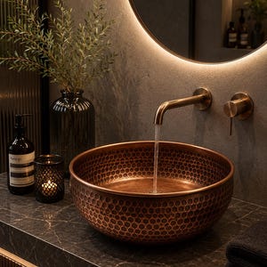 Puede incluir: Un lavabo redondo de cobre martillado con un patrón de panal texturizado en el exterior. Un grifo de pared de bronce cepillado dispensa un flujo constante de agua hacia el cuenco. El lavabo está colocado sobre una encimera de piedra oscura veteada.