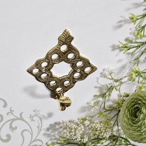 May include: A gold-toned metal wall hook featuring a diamond-shaped backplate with an intricate openwork lattice pattern and a pointed top. The hook extends from the bottom of the decorative plate, which is finished with a polished metallic sheen.