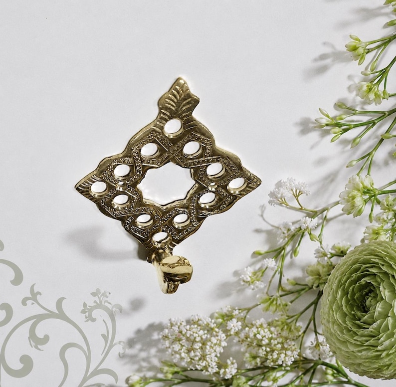 Pu&ograve; includere: Un gancio da parete in metallo color oro con una piastra posteriore a forma di diamante, caratterizzata da un intricato motivo a reticolo traforato e una punta superiore. Il gancio si estende dalla base della piastra decorativa lucida.