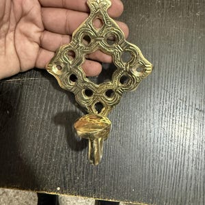 Pu&ograve; includere: Un'applique da parete in ottone lucido caratterizzata da un'intricata piastra posteriore traforata a forma di diamante con incisioni dettagliate e un'apertura circolare centrale. Una piccola coppa portacandela sporge dalla struttura.