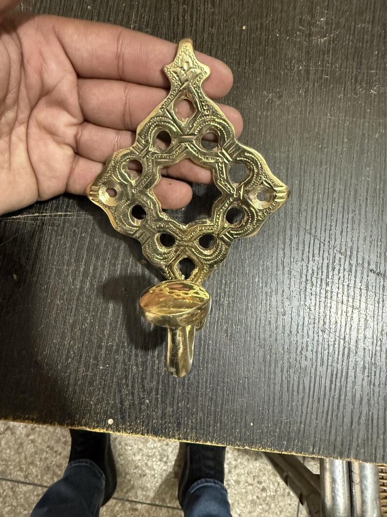 Pu&ograve; includere: Un'applique da parete in ottone lucido caratterizzata da un'intricata piastra posteriore traforata a forma di diamante con incisioni dettagliate e un'apertura circolare centrale. Una piccola coppa portacandela sporge dalla struttura.