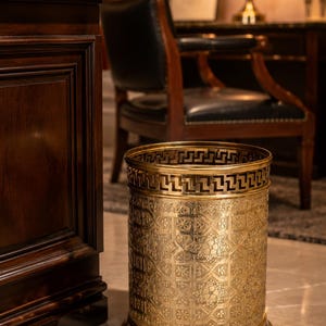 May include: A cylindrical brass wastebasket featuring an intricate embossed geometric pattern across its body and a decorative Greek key motif along the upper rim. The metal surface has a polished gold-toned finish with a wide, stable base.