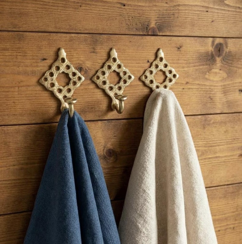 Pu&ograve; includere: Tre ganci da parete in metallo color oro con un design traforato a forma di diamante. Due dei ganci sostengono asciugamani in tessuto, uno di colore blu scuro tinta unita e l'altro di colore beige chiaro tinta unita, mentre il terzo resta vuoto.