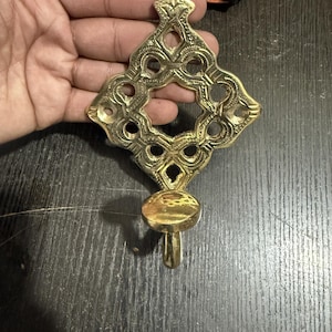Pu&ograve; includere: Un gancio da parete in ottone lucido caratterizzato da una piastra posteriore decorata a forma di diamante, con intricati motivi intagliati e un gancio arrotondato alla base. La superficie metallica &egrave; riflettente e color oro.