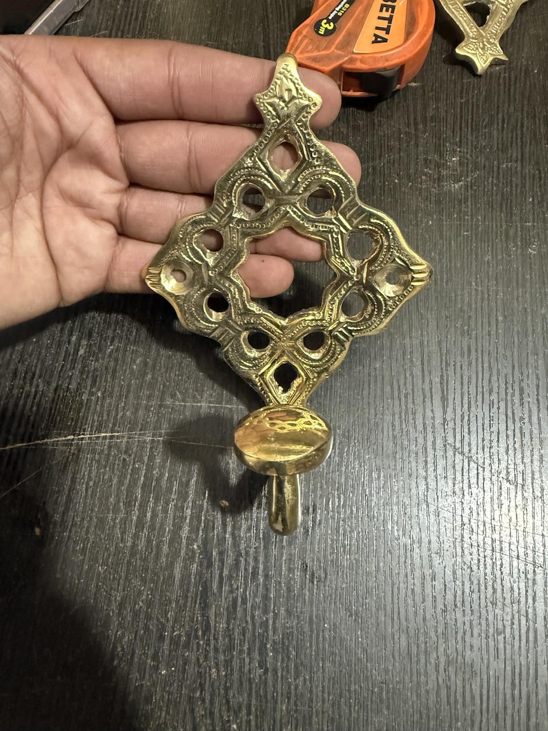 Pu&ograve; includere: Un gancio da parete in ottone lucido caratterizzato da una piastra posteriore decorata a forma di diamante, con intricati motivi intagliati e un gancio arrotondato alla base. La superficie metallica &egrave; riflettente e color oro.