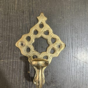 Pu&ograve; includere: Un gancio da parete in ottone caratterizzato da una piastra posteriore a forma di diamante traforato con motivi incisi decorativi e un ritaglio circolare centrale. Il gancio si estende verso l'esterno dalla parte inferiore della piastra metallica.