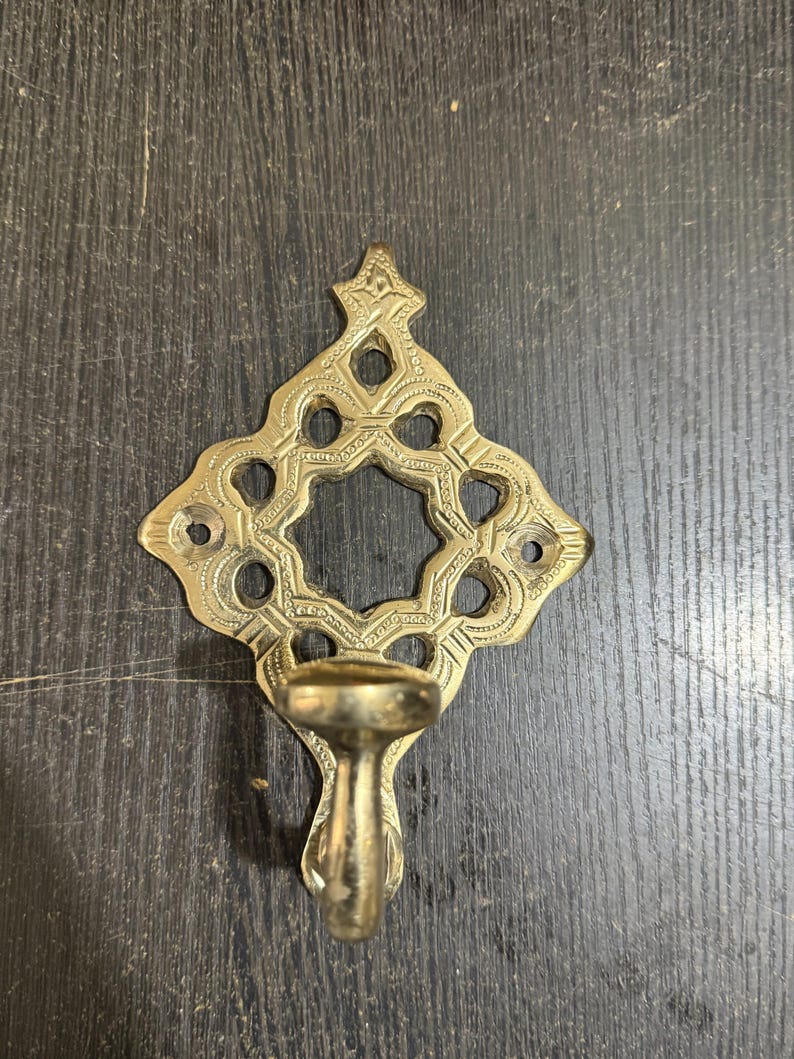 Pu&ograve; includere: Un gancio da parete in ottone caratterizzato da una piastra posteriore a forma di diamante traforato con motivi incisi decorativi e un ritaglio circolare centrale. Il gancio si estende verso l'esterno dalla parte inferiore della piastra metallica.